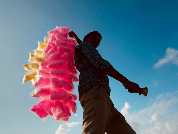 Silhouette of a street vendor carrying colorful cotton candy bags under a bright blue sky in Asia.