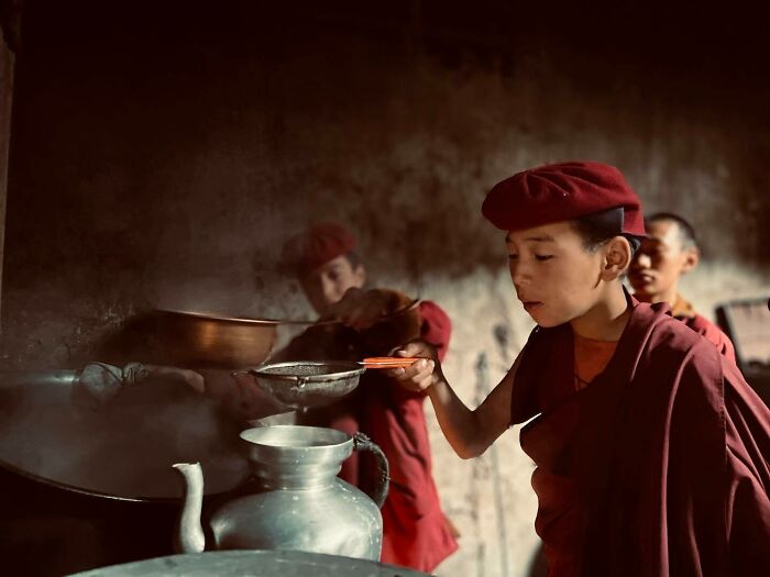 Young monk in red robes and hat cooking in a rustic Asian kitchen, showcasing authentic cultural photography from Asia.