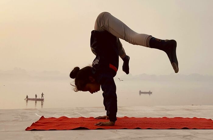 Person practicing yoga handstand on red mat near calm water with boats, reflecting the spirit of exploring Asia through photography.