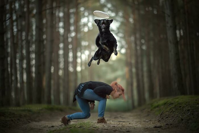 Black dog flying through the air like a superhero while a person crouches in a forest path on a cloudy day.