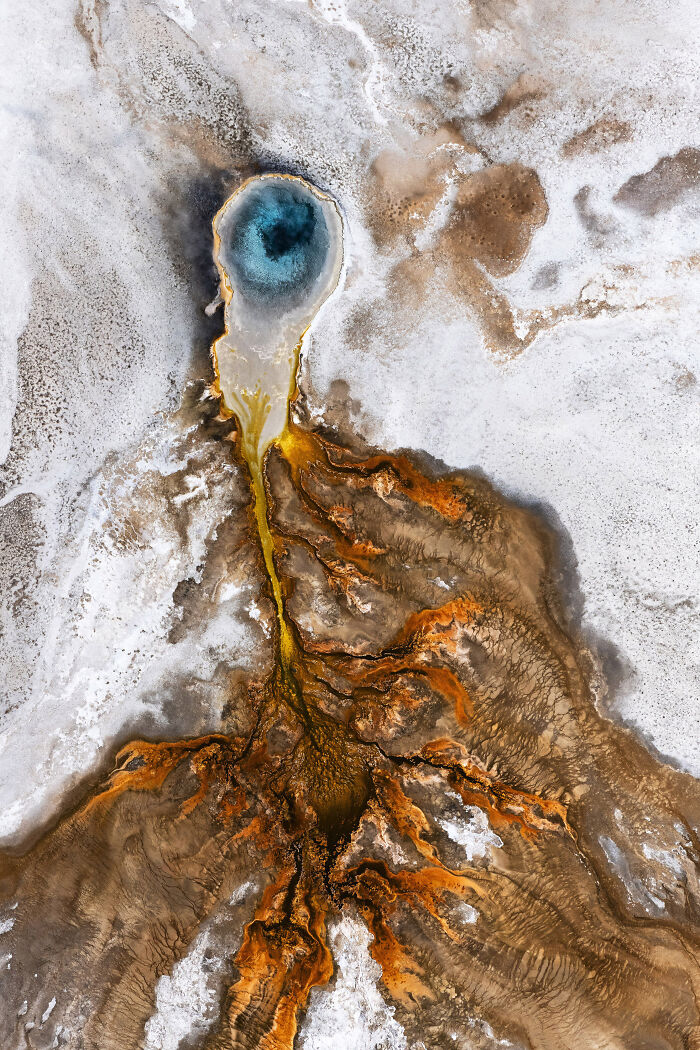 Aerial shot of a colorful geothermal pool with orange and brown mineral deposits in a barren landscape.