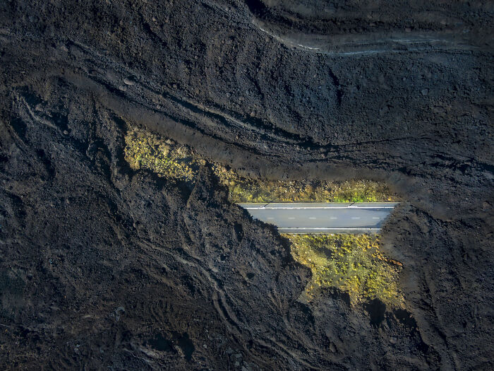 Aerial shot of a road cutting through rugged dark volcanic terrain with patches of green vegetation.