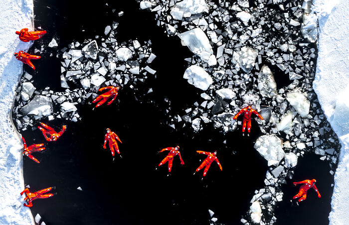 Aerial shot of people in red suits floating on dark icy water surrounded by broken ice chunks.