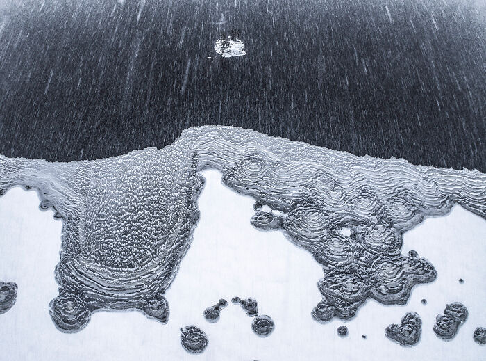 Aerial shot of icy river patterns with snow and rain falling, showcasing nature’s texture and contrast in a winning photography style.