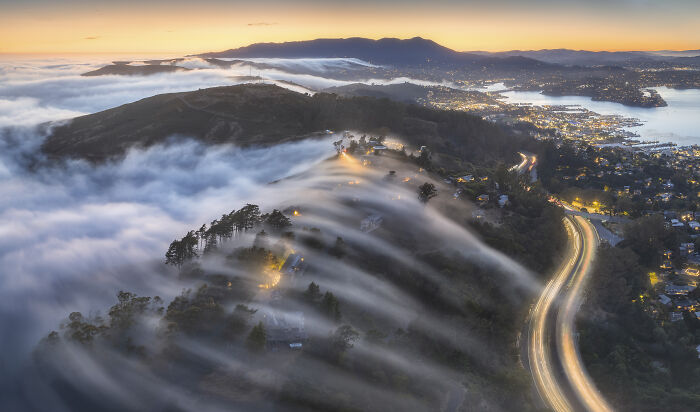 Aerial shot of hillside town with fog rolling over trees and winding illuminated highway at sunset, showcasing stunning landscape views.