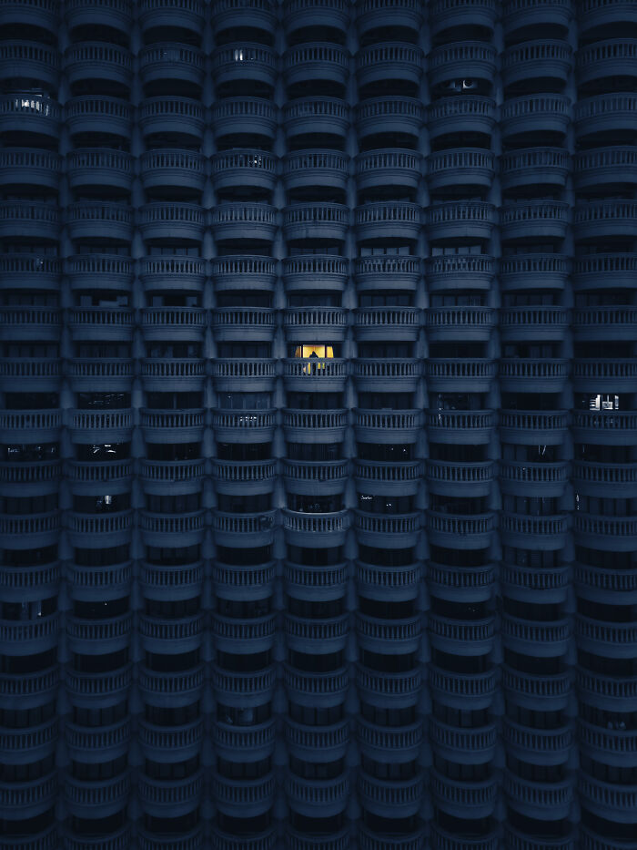 Rows of dark balconies at night with one glowing window showing silhouettes in winning aerial shot.