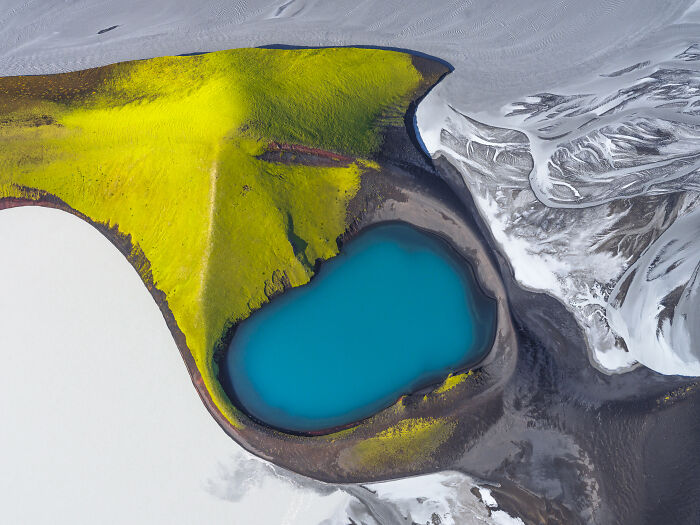 Aerial shot of a vibrant blue lake surrounded by yellow-green vegetation and snowy white landscape.