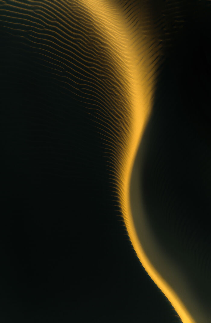 Aerial shot of sand dunes with golden light highlighting ripples and smooth curves in a dark desert landscape.