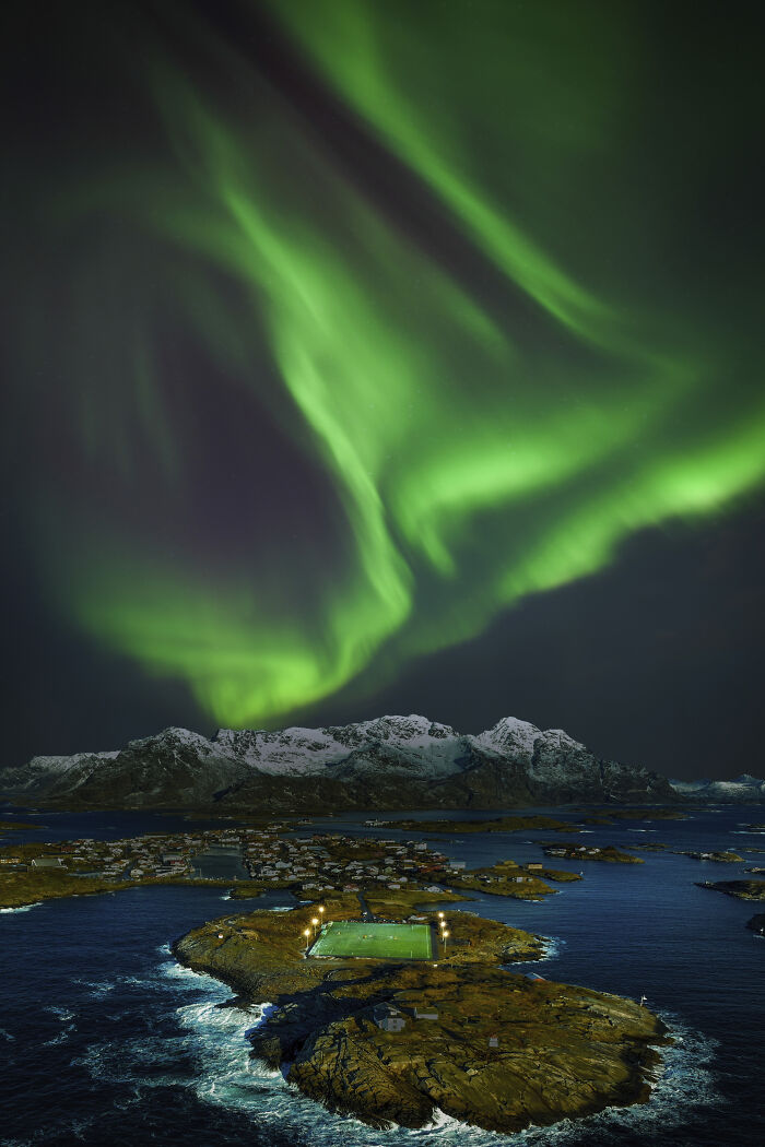 Aerial view of a small island with a lit soccer field under vibrant green northern lights at night.