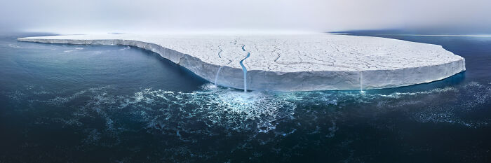 Aerial shot of a massive ice shelf with a waterfall flowing into the dark ocean, showcasing stunning natural contrasts.