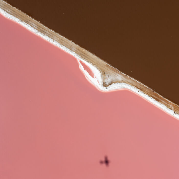 Aerial shot of contrasting pink and brown salt ponds divided by a narrow textured land strip with a small airplane shadow below.