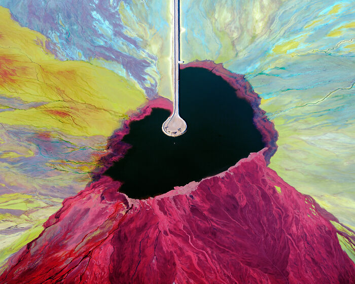 Aerial shot of a vibrant red and yellow landscape with a dark central pool and a vertical structure reaching into it.