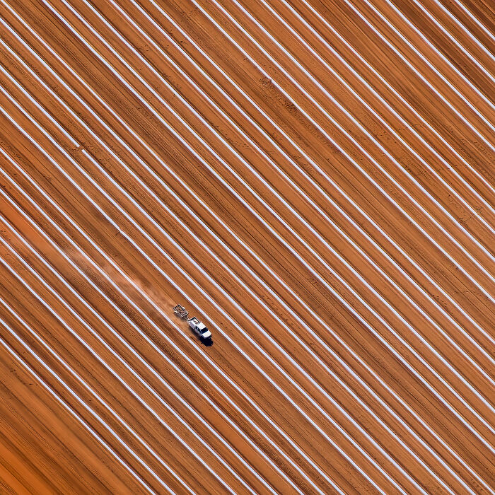Aerial shot of a vehicle driving through long parallel rows in a reddish agricultural field, showcasing patterns and textures.