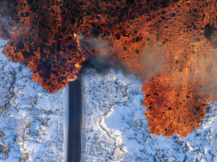 Aerial shot capturing molten lava flowing beside a snowy landscape and a road, showcasing striking natural contrasts.