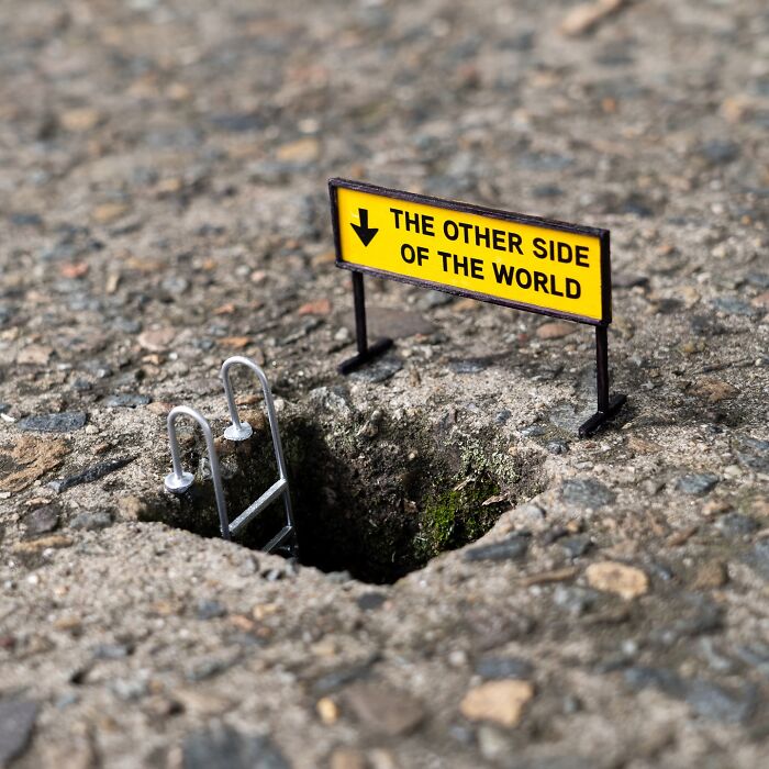 Tiny street scene with a miniature ladder and a yellow sign reading THE OTHER SIDE OF THE WORLD on asphalt.