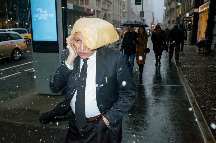 Man in suit shielding head with plastic bag while walking through rainy NYC street, capturing striking street photos and city stories.