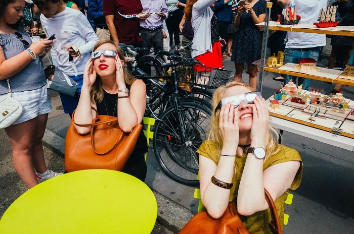 Two women in NYC street scene sitting and looking up wearing eclipse glasses, surrounded by bicycles and pedestrians.