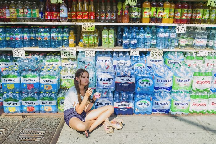 Young woman eating ice cream sitting on sidewalk in front of bottled water display in a powerful street moment of NYC.