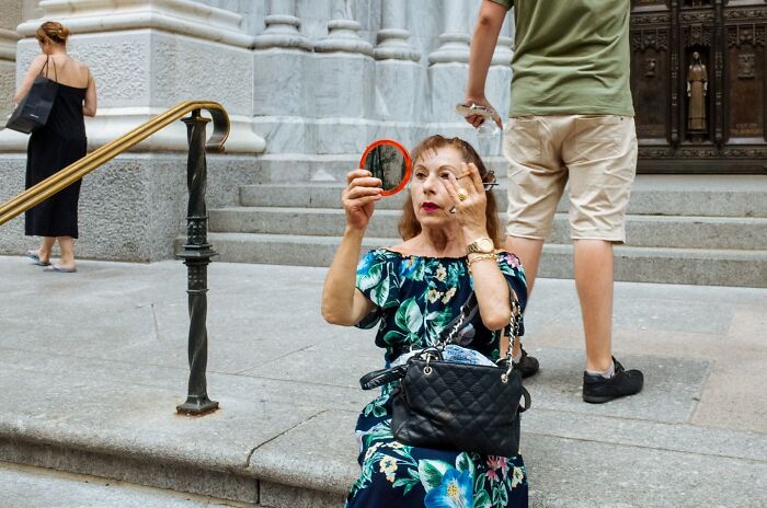 Woman in floral dress sitting on NYC street steps, applying makeup with a handheld mirror in vibrant street photo.