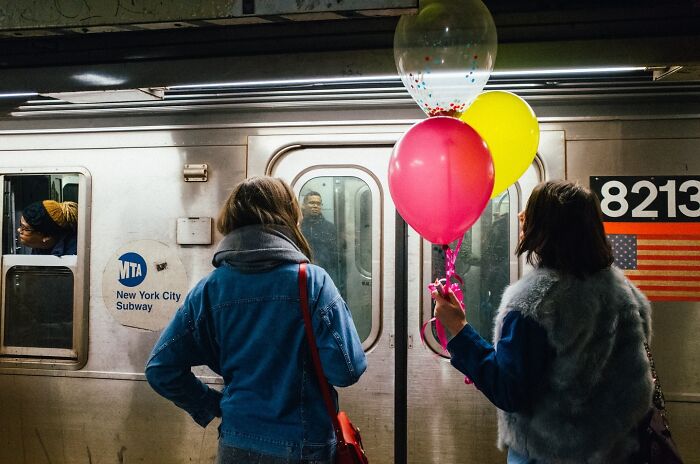 Two women holding colorful balloons wait on a platform as a New York City subway train arrives.