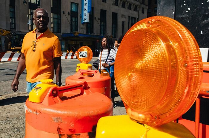 Man in orange shirt walking near street construction barrels in a powerful street moment captured in NYC.