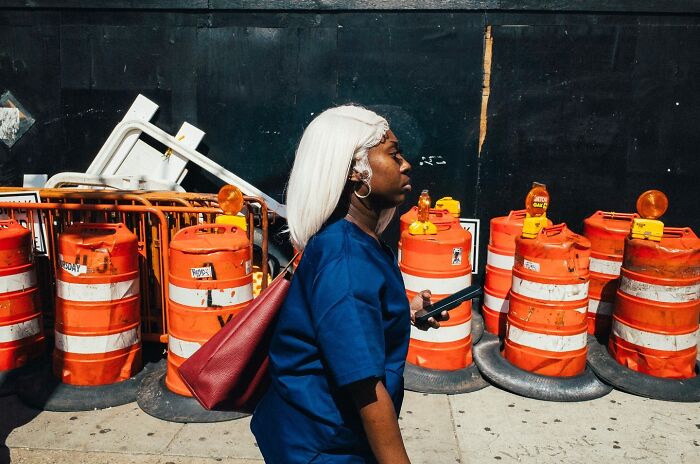 Woman with white hair walking past orange construction barrels in a striking NYC street photo capturing city stories.