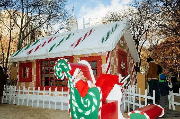 Woman dressed as Santa in a festive chair in front of a holiday gingerbread house in a street moment of NYC.
