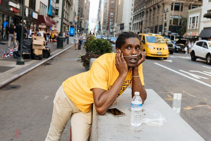 Man in yellow shirt leaned on a street barrier in NYC with a water bottle and oxygen tube, captured in striking street photos.