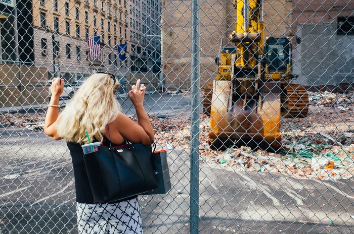 Woman with blonde hair and black bag standing by fence near construction site, capturing powerful street moments in NYC.