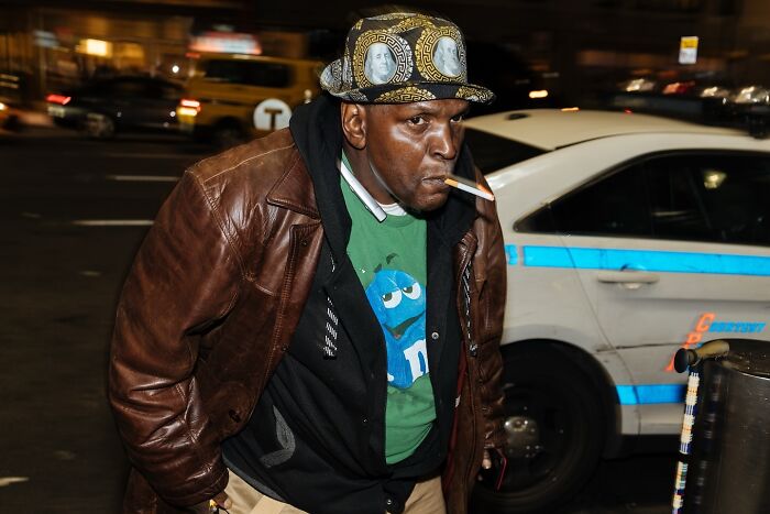 Man wearing stylish hat and leather jacket smoking cigarette on busy NYC street at night, capturing powerful street moments.
