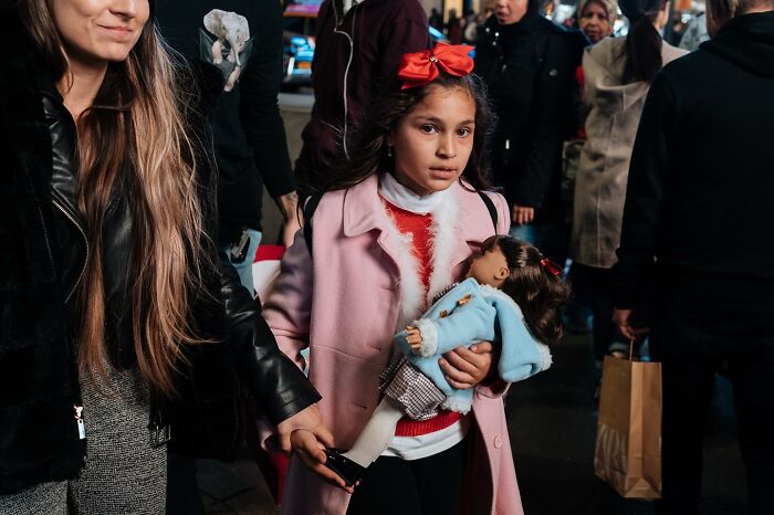 Young girl in a pink coat holding a doll while walking through a busy NYC street captured in powerful street moments photography