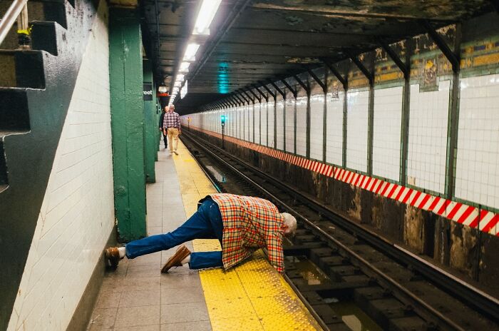 Man in a plaid jacket leaning over subway tracks at NYC station, a striking street photo capturing urban life moments.