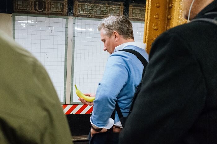 Man in blue shirt holding a banana on a NYC subway platform, a striking street photo capturing city life moments.