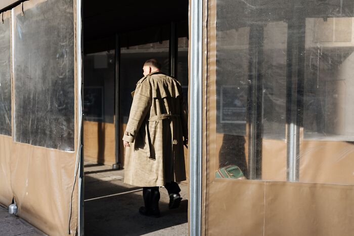 Man in a long coat walking between plastic-covered outdoor seating, capturing powerful street moments of NYC life.