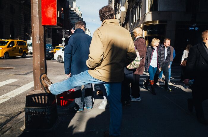 Man stretching leg on busy NYC street corner with pedestrians and yellow cabs capturing street moments in NYC.