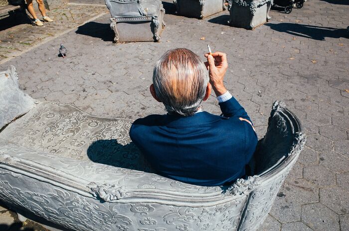 Man in blue suit smoking cigarette seated on ornate bench in NYC street, captured in striking street photo by Mathias Wasik