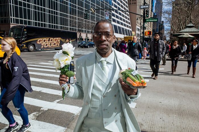 Man in light suit holding flowers and vegetables on a busy NYC street, capturing powerful street moments of NYC.