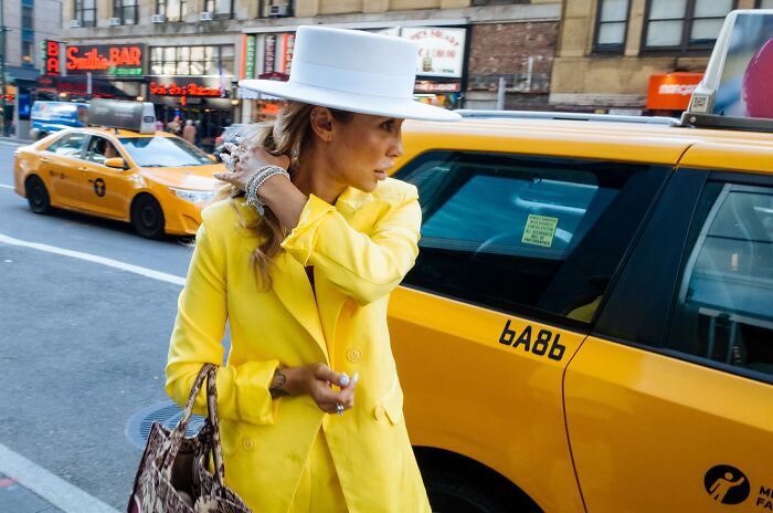 Woman in yellow coat and white hat standing by a NYC yellow taxi in a striking street photo capturing city stories.