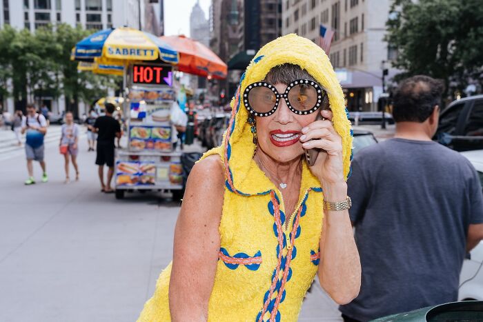 Woman in bright yellow hoodie and oversized sunglasses talking on phone on busy NYC street, capturing striking street photos.