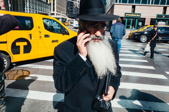 Street photographer captures powerful NYC moment with bearded man in black hat talking on phone near yellow taxi.