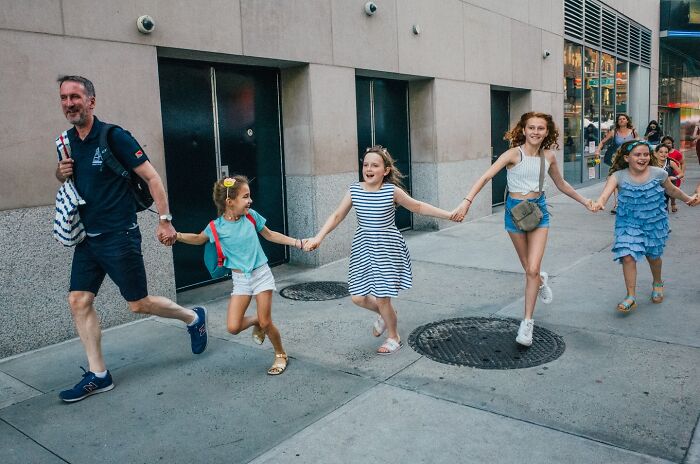 Man and four girls holding hands, running joyfully on a city sidewalk, capturing powerful street moments of NYC.