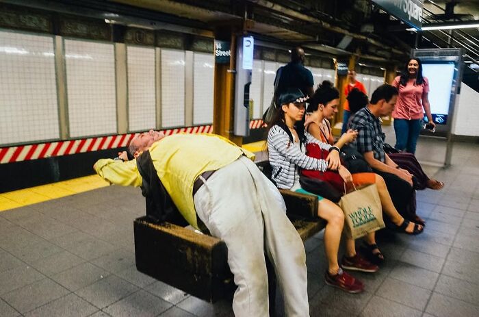 Man in yellow shirt resting on bench while others wait on subway platform in NYC street photo capturing city stories by Mathias Wasik