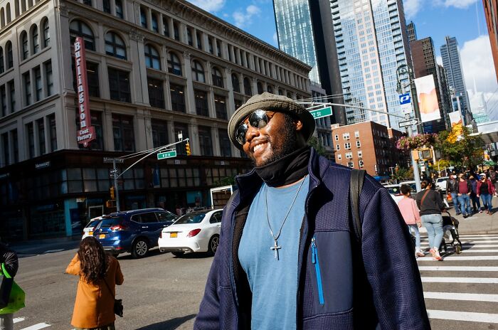 Smiling man wearing sunglasses and a hat walking on a busy NYC street captured in powerful street moments photography.