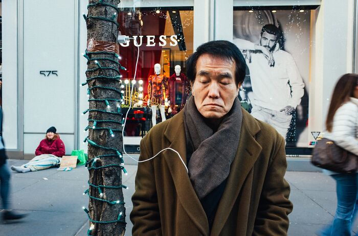 Man in brown coat with scarf stands near tree wrapped in lights on busy NYC street in a candid street photo.