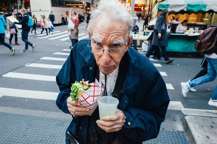 Elderly man eating street food with drink at busy NYC crosswalk, showcasing striking street photos of city life.