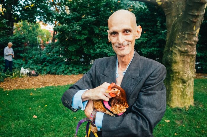 Man in a suit holding a chicken in a green park, captured in a striking NYC street photo by Mathias Wasik.