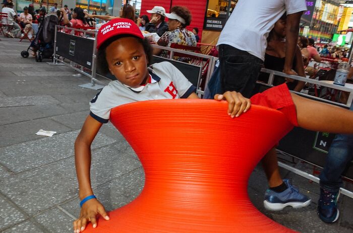 Young boy in a red hat spinning on an orange street chair among crowds in a lively NYC street moment captured by a photographer.