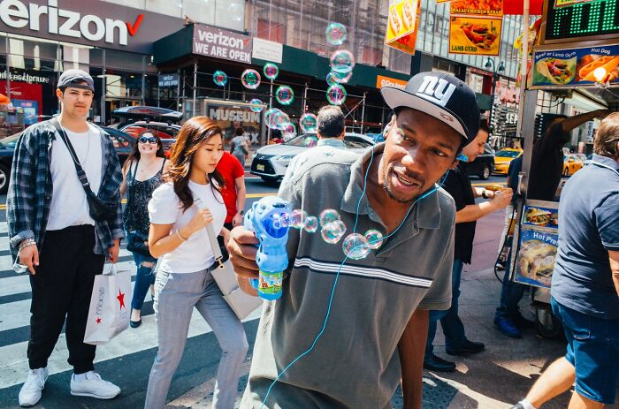 Man wearing a NY cap blowing bubbles on a busy NYC street filled with pedestrians and vibrant city life street photos.