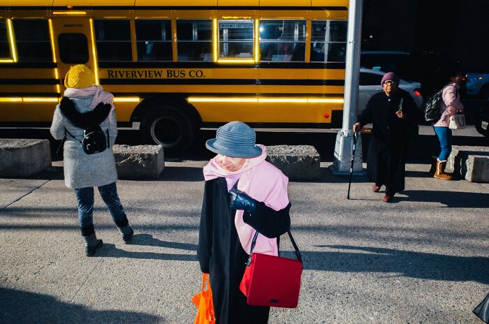 Woman with red bag and blue hat walking near a yellow school bus in a striking NYC street photo.