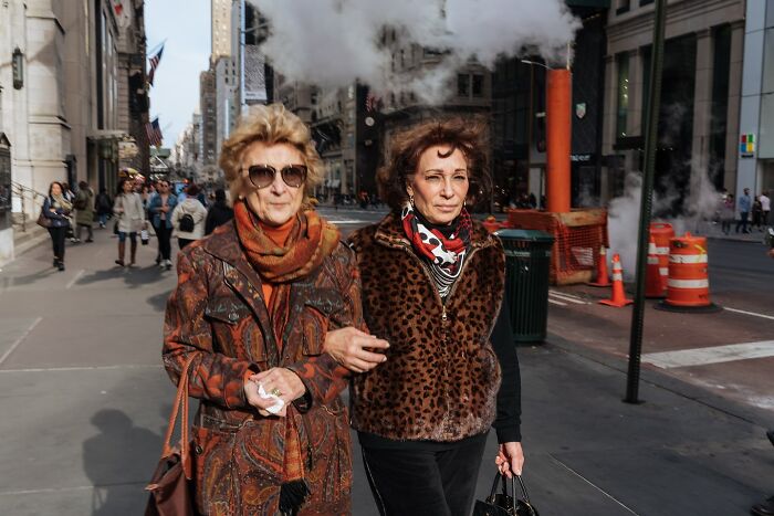 Two women walking on a busy NYC street with steam rising, capturing the essence of striking street photos in New York City.